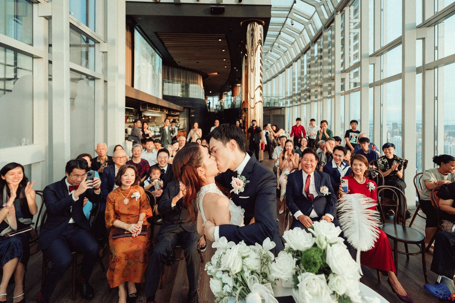 Wedding guests gathered in a modern venue with floor-to-ceiling windows and white floral decorations.