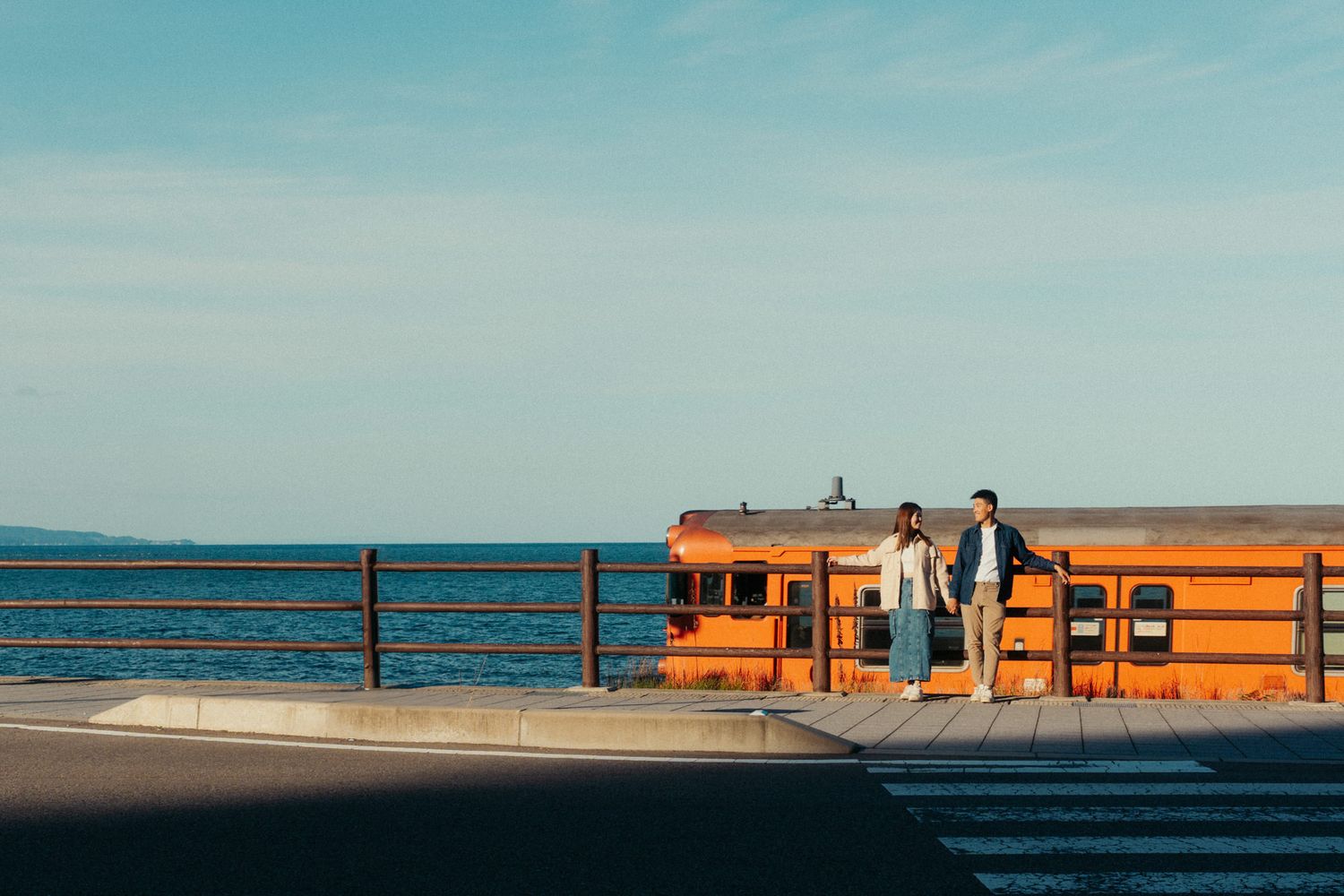 Orange train cars sit against a bright blue sky on a coastal railway line.
