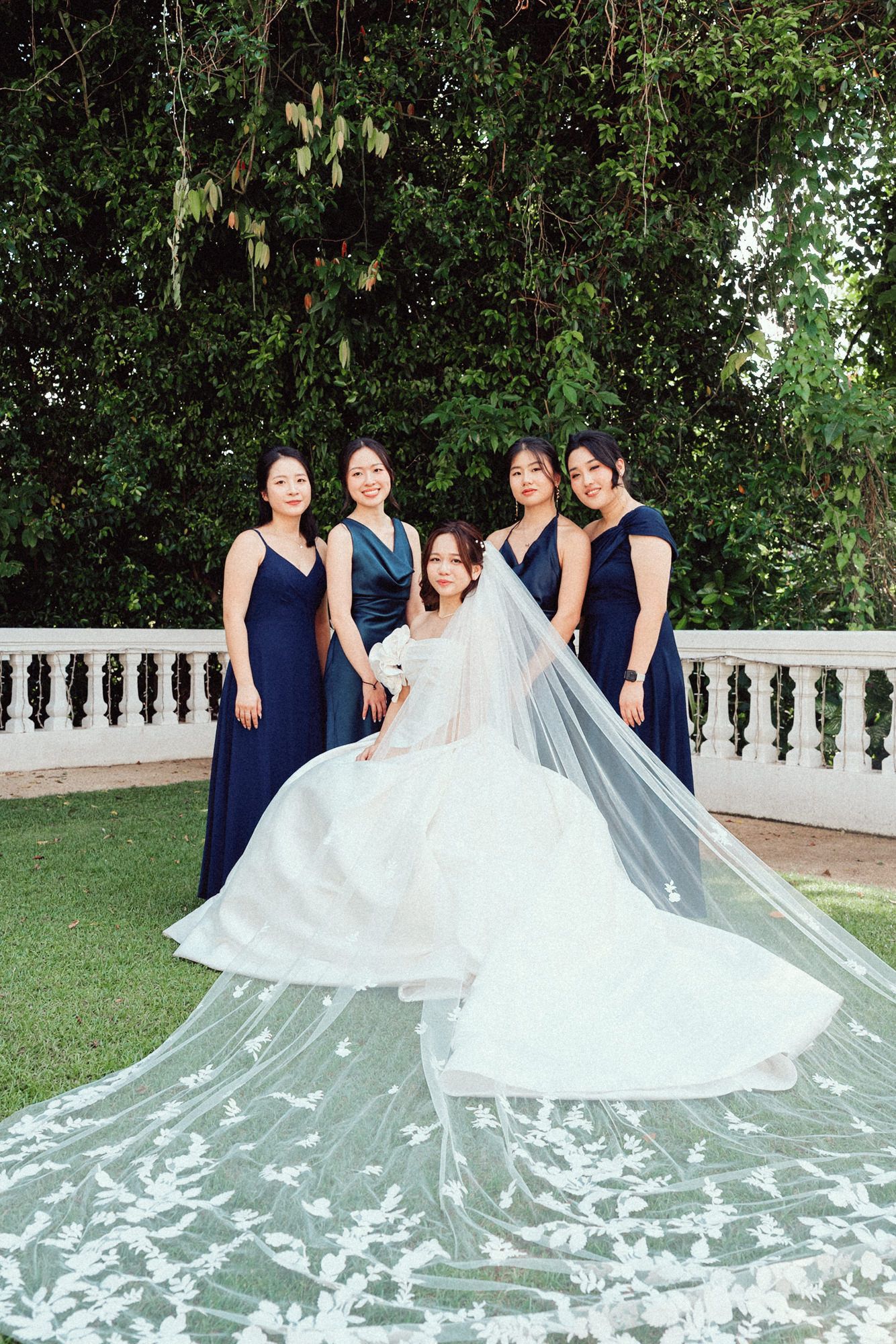 A bridal party in navy blue dresses poses with a bride in a flowing white gown with a dramatic cathedral-length veil outdoors.
