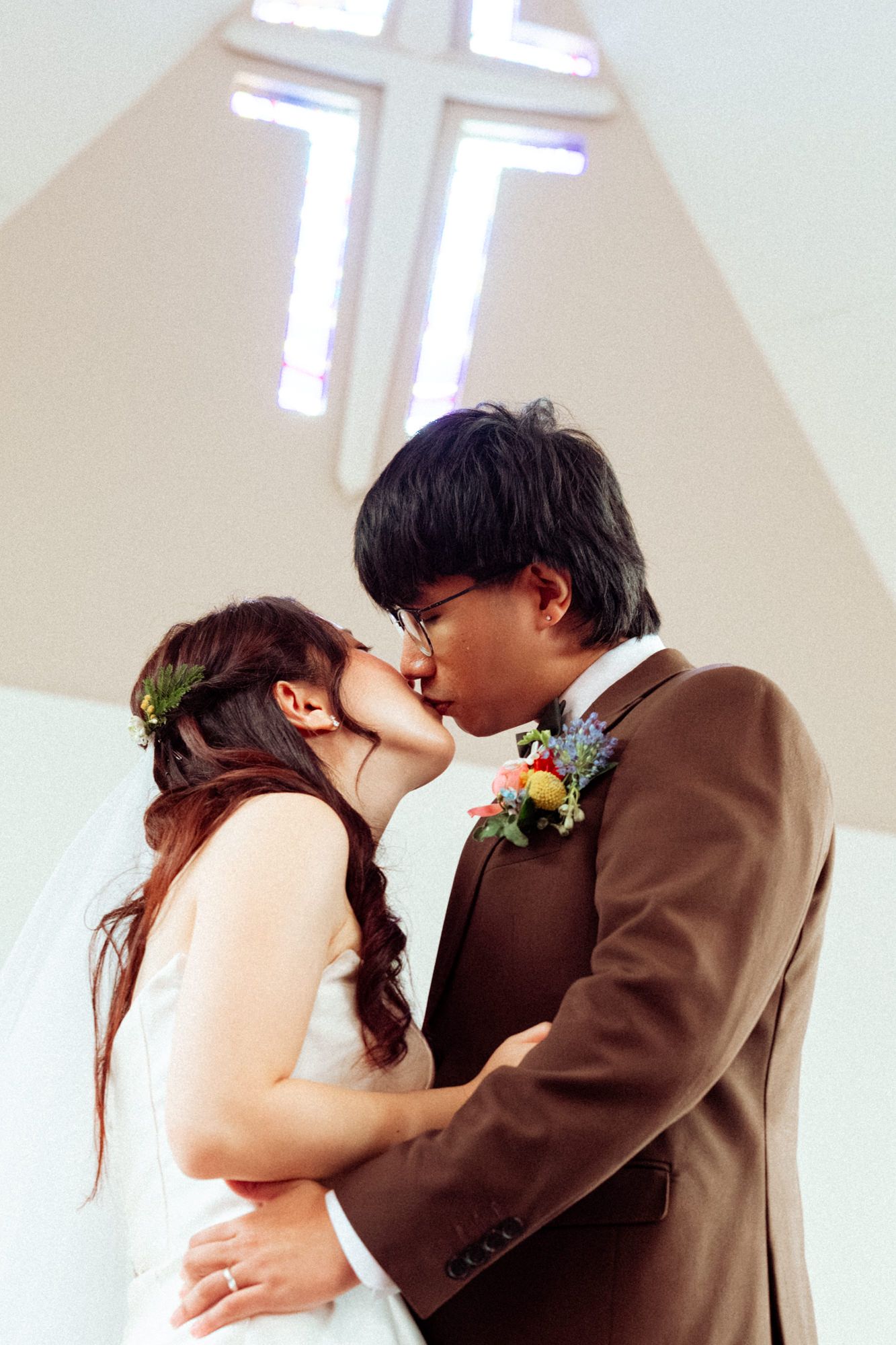 Bride and groom sharing first kiss at church altar with glowing cross window backdrop in wedding ceremony.
