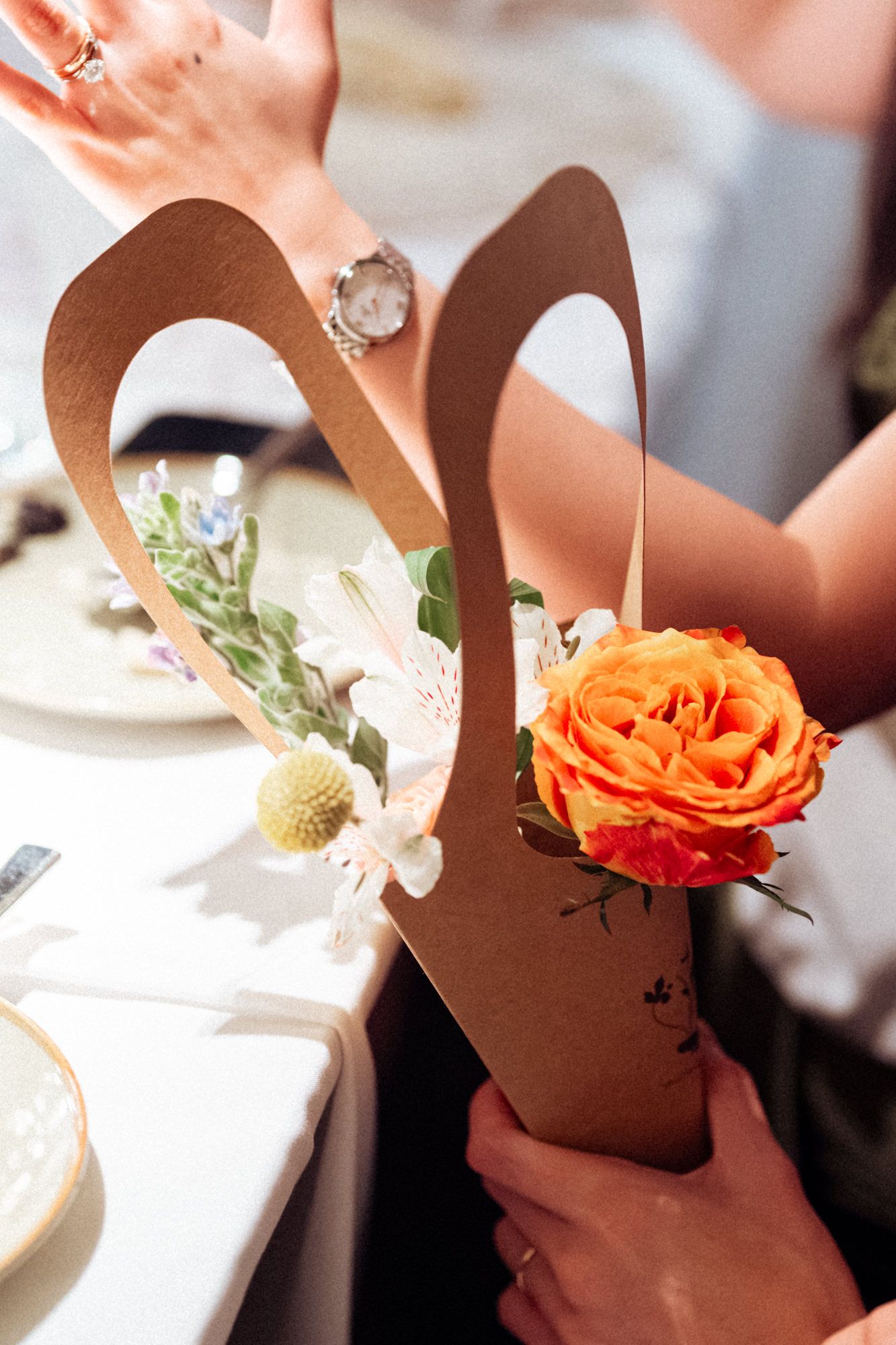Hands holding a kraft paper cone bouquet with an orange rose and white flowers at an elegant event.