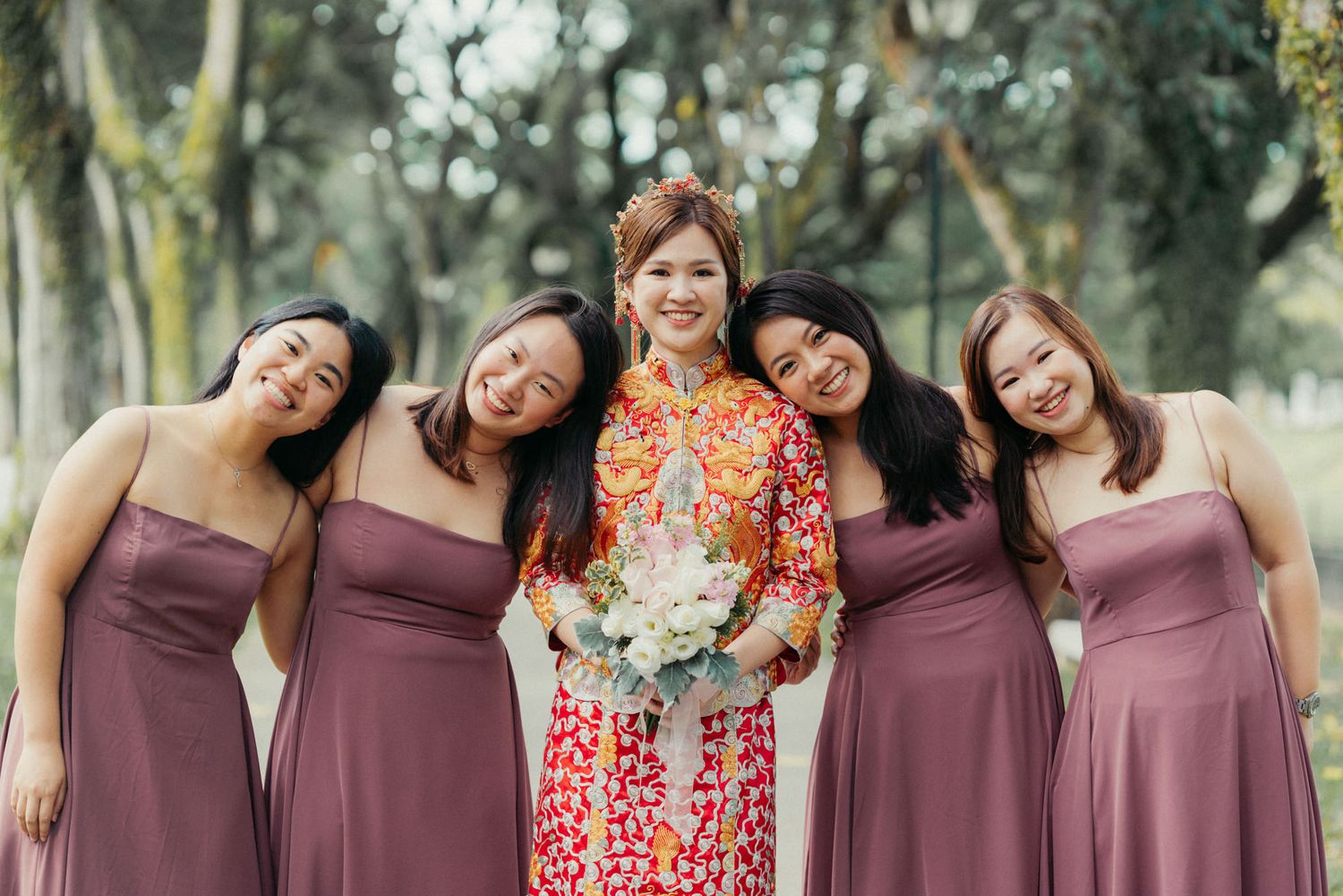 Bride in colorful ao dai holds bouquet surrounded by four bridesmaids in mauve dresses outdoors.