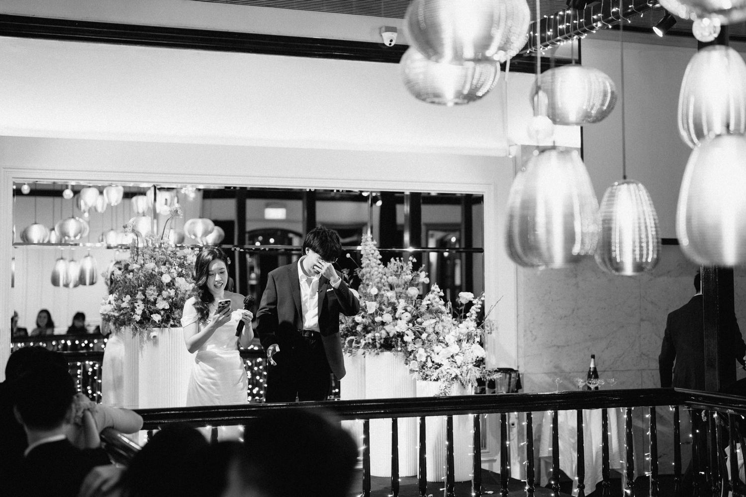 Black and white photo of bride and groom standing at wedding reception altar surrounded by floral arrangements and hanging lanterns.