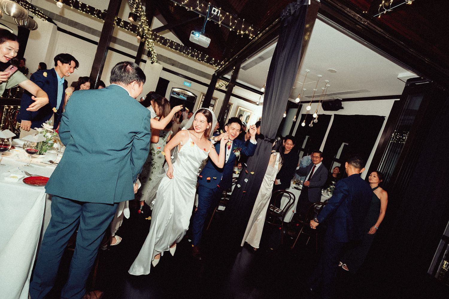 People dancing and celebrating at a wedding reception in a dimly lit venue with festive decorations.