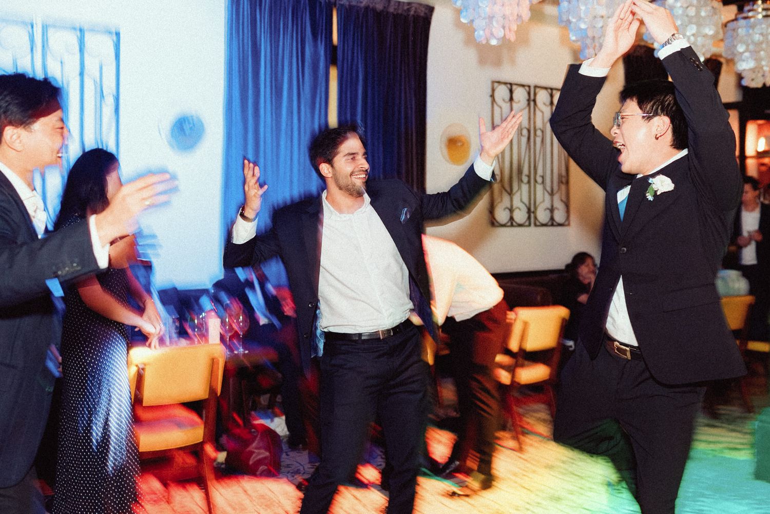 People dancing and celebrating at a party with colorful lighting and blue curtains in the background.
