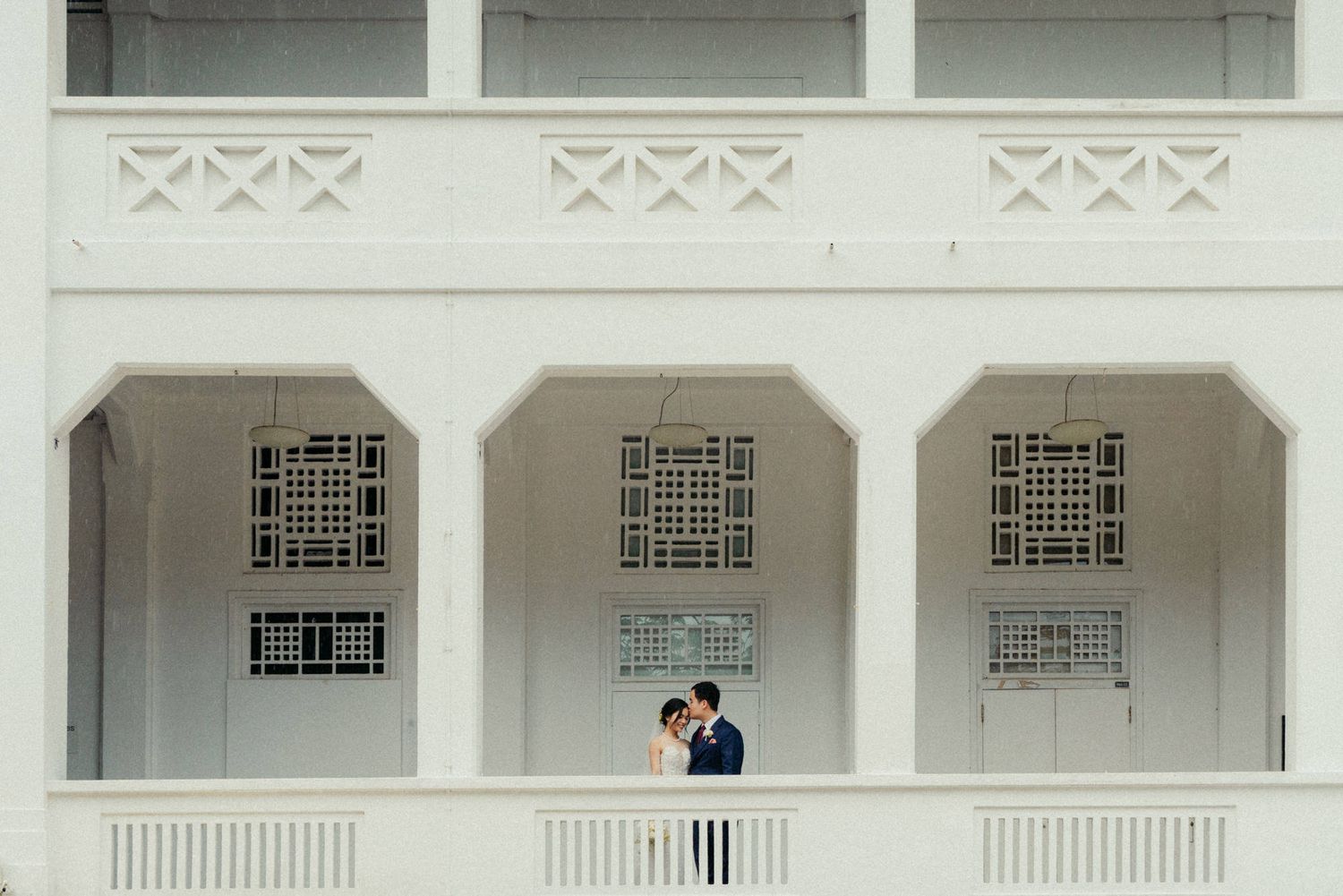 Couple embracing on white colonial building balcony, elegant arched architecture framing romantic wedding portrait.