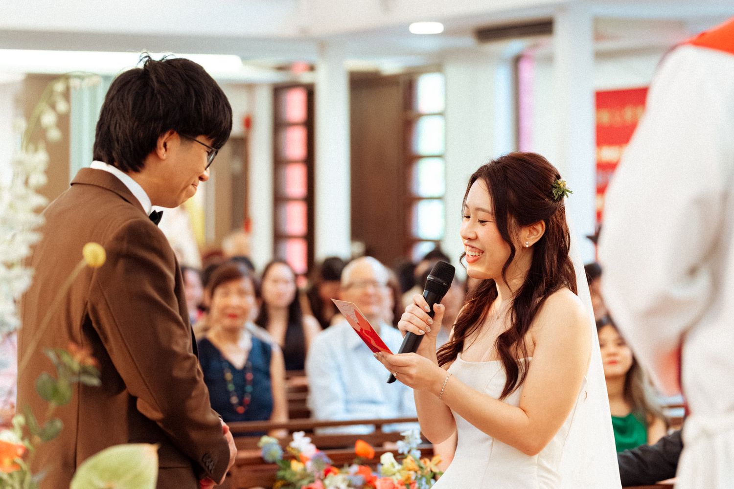Bride smiles while reading vows into a microphone at a church wedding ceremony, with groom in brown suit facing her.