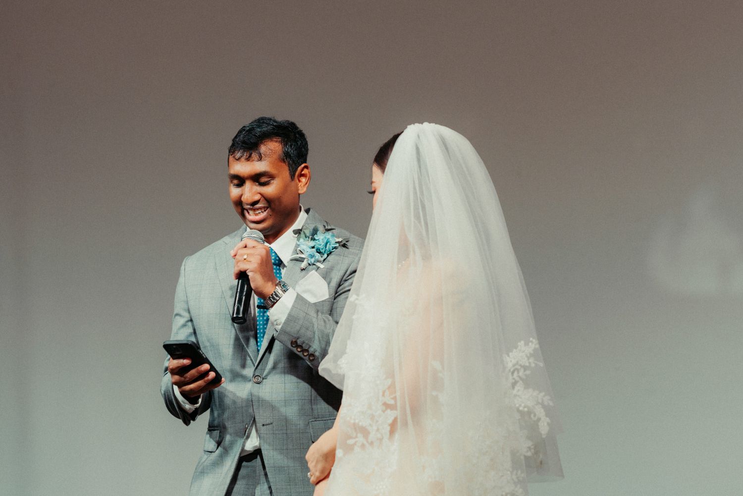 Groom in gray suit reads vows from phone while laughing with bride in white wedding dress and veil at ceremony.