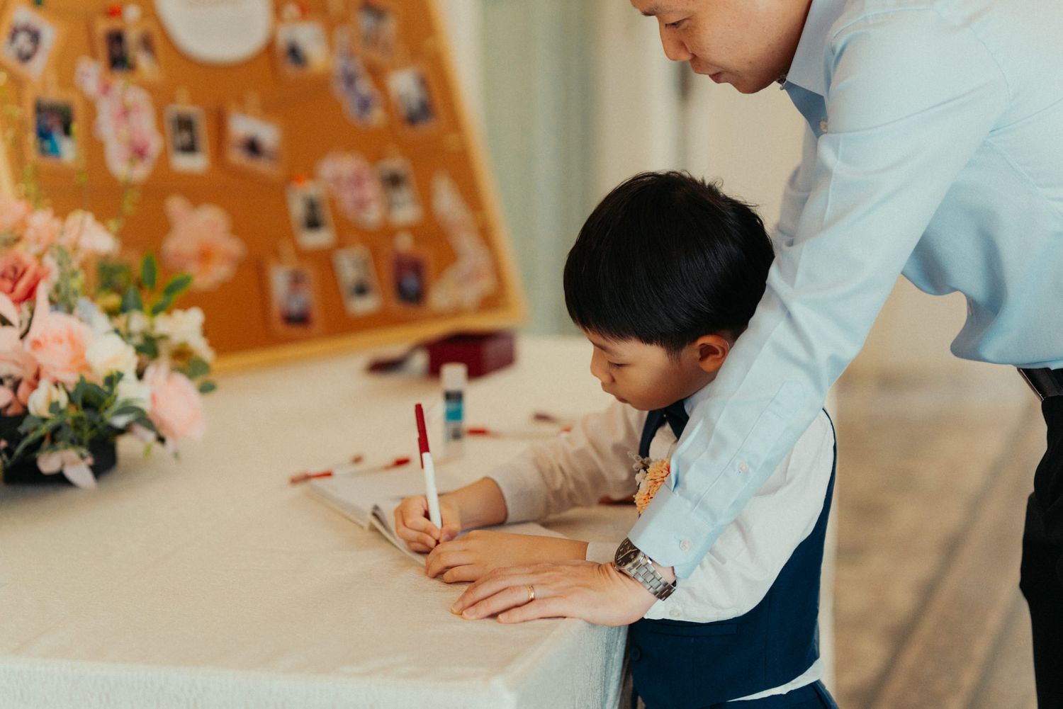 Adult guiding a young child writing at a table during a wedding reception event.