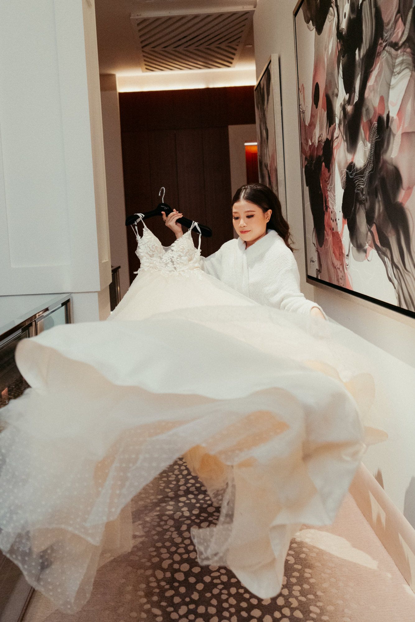 Bride in white robe holding elegant lace wedding gown on hanger in luxury hotel corridor with abstract art.