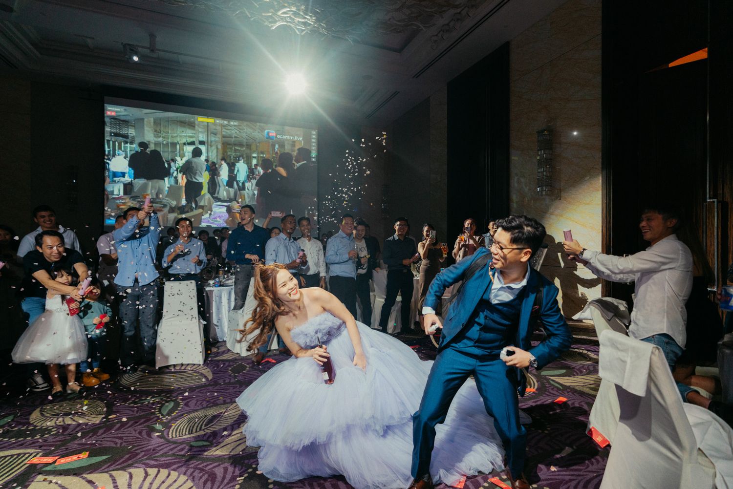 Bride and groom dancing joyfully at wedding reception with guests celebrating and confetti flying.