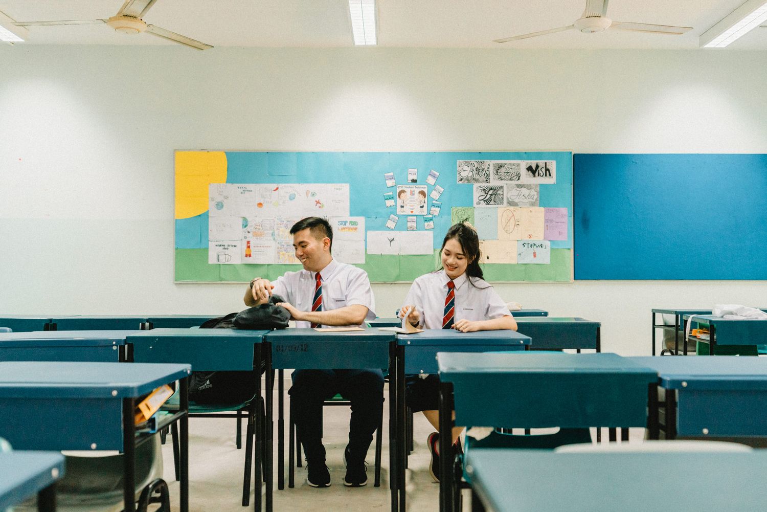 Students sitting at desks in a bright classroom with blue walls and ceiling fans.