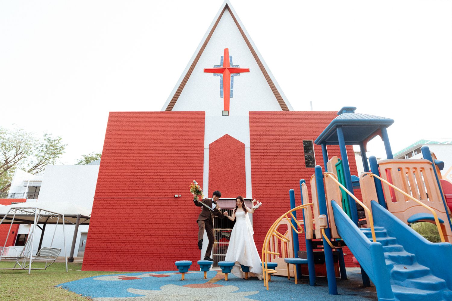 Couple celebrating in front of a red and white church with a cross, near a colorful playground outside.