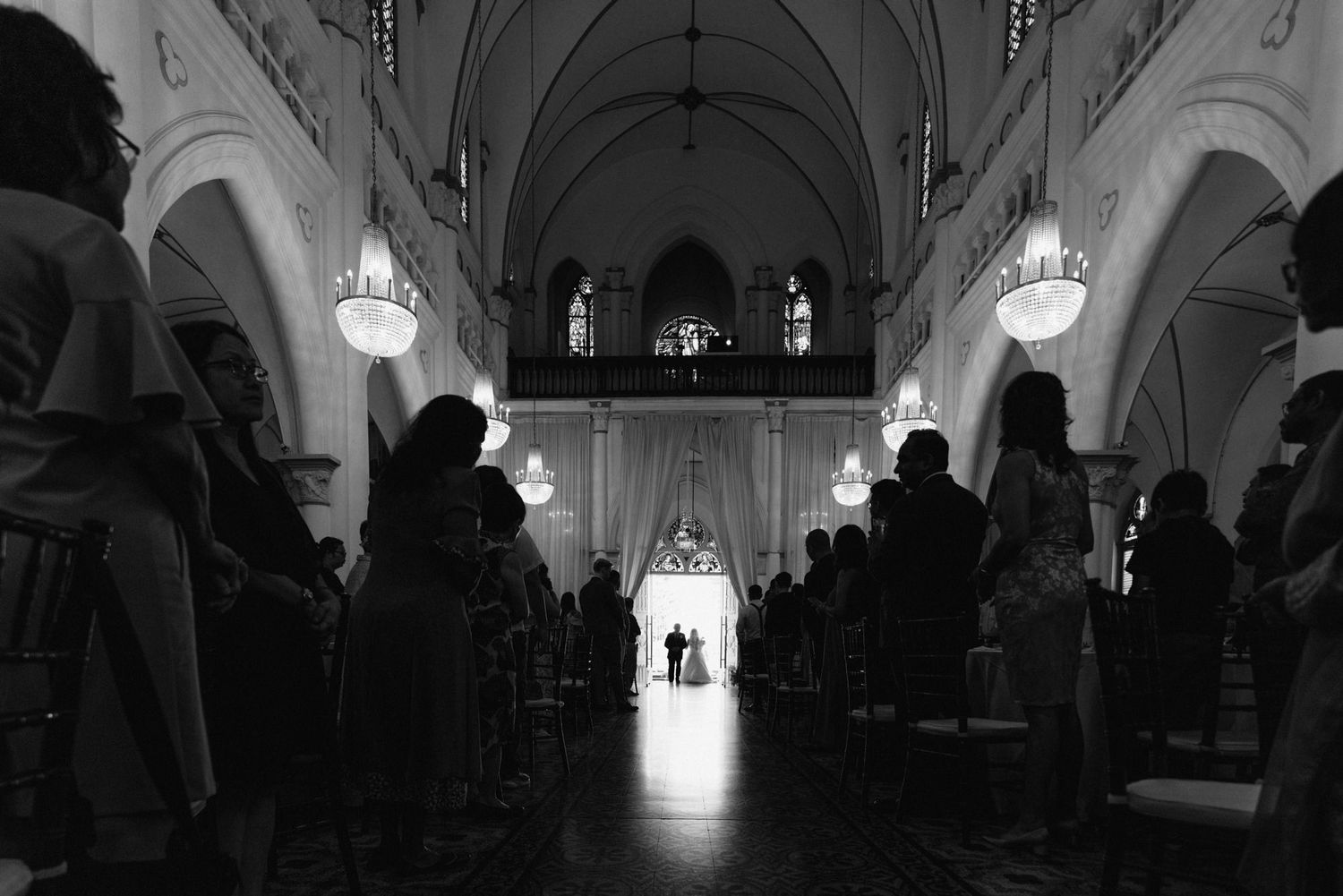 Black and white photo of a wedding ceremony inside a grand church with chandeliers and guests seated in pews.