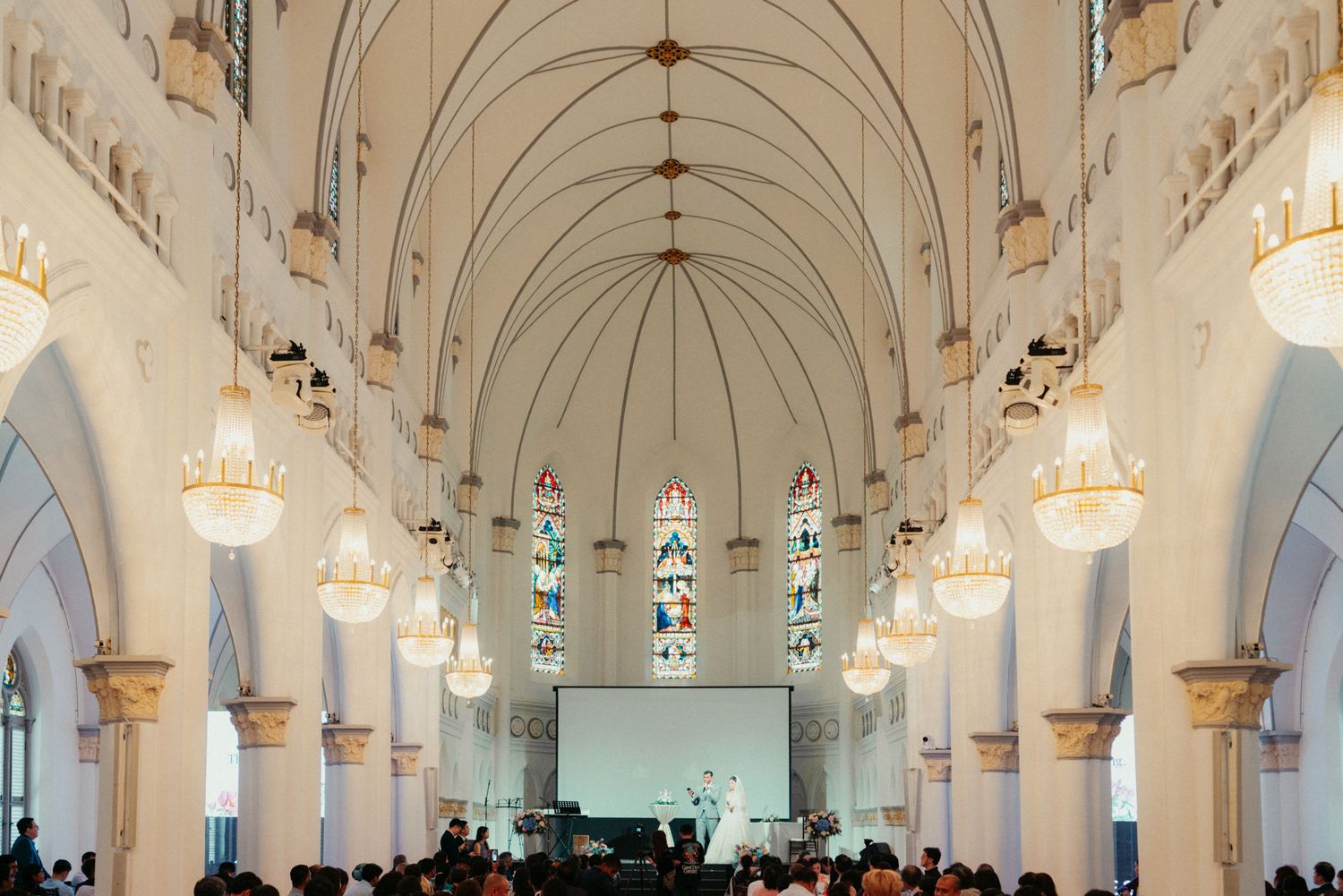 Grand white cathedral interior with vaulted ceilings, stained glass windows, chandeliers, and audience gathered for a presentation.
