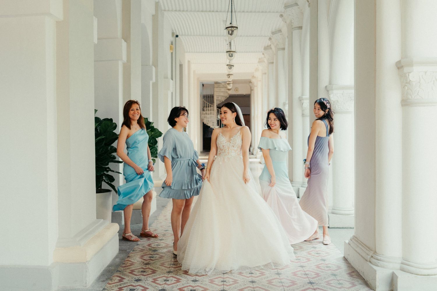 Bride in white ball gown walks with bridesmaids in blue and purple dresses along elegant colonial corridor.