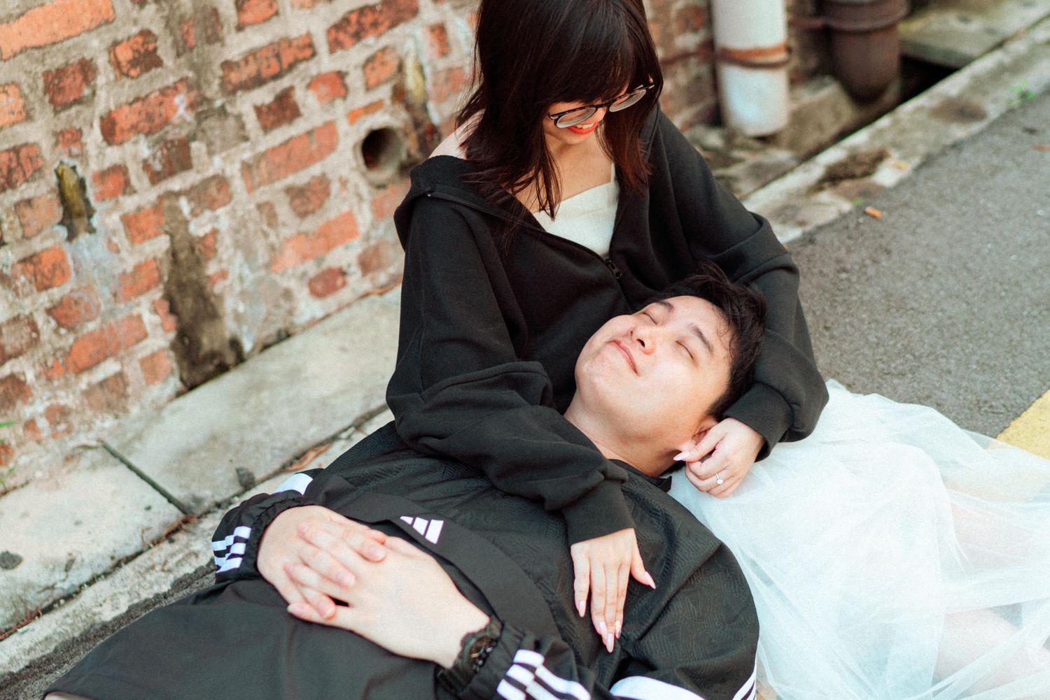 A couple in black clothing lies together on concrete ground near a brick wall.