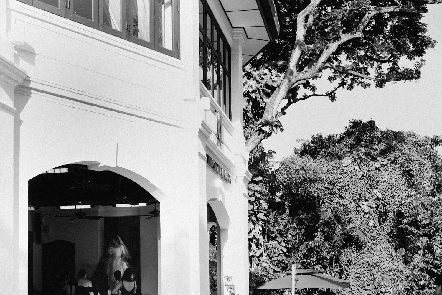 Black and white architectural photograph showing an arched entrance to a building with lush tropical foliage visible.