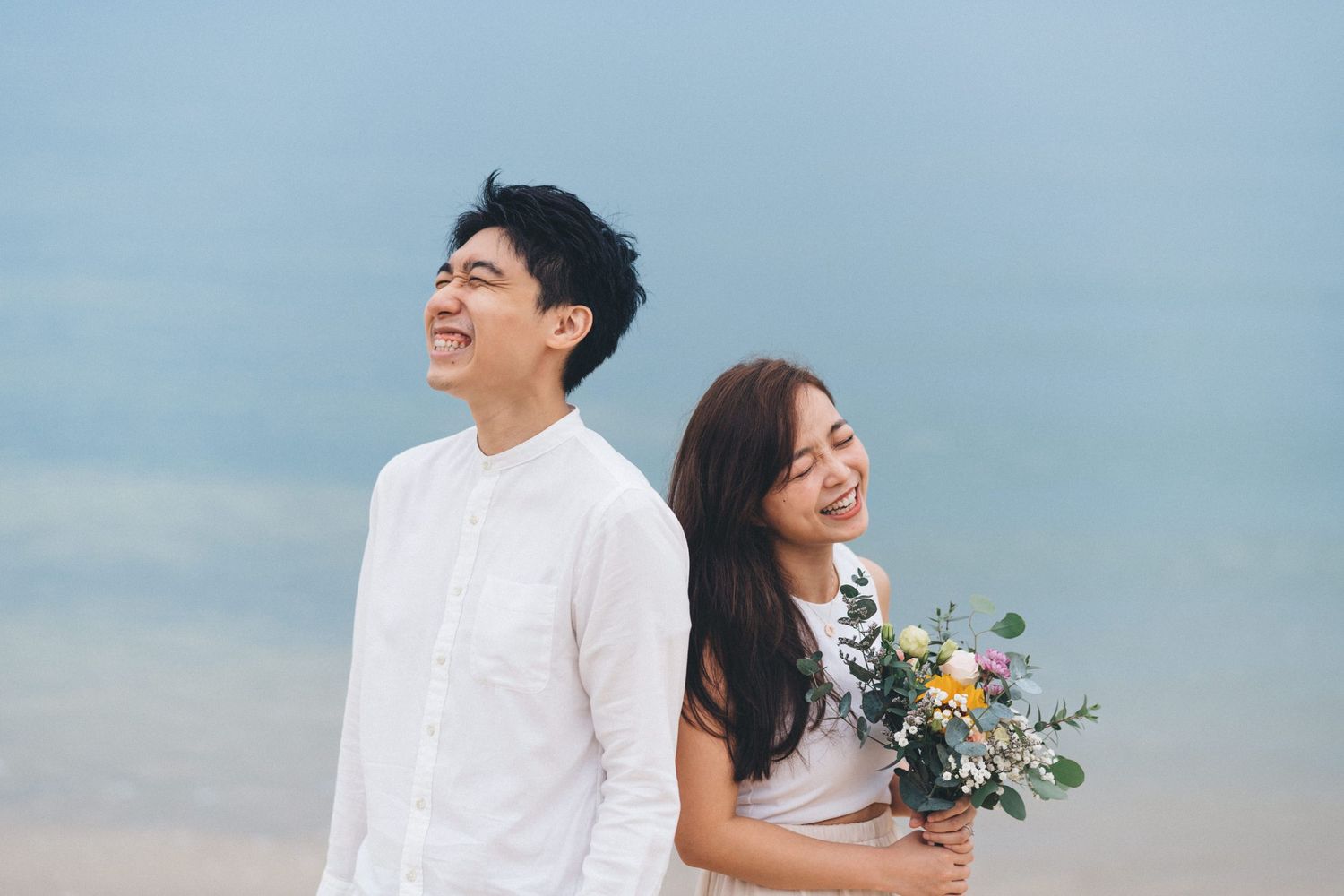A couple in white stands against a blue sky backdrop while holding a bouquet of flowers and greenery.