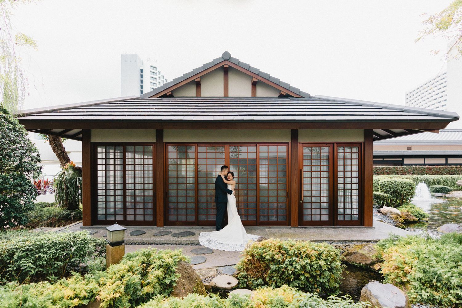 Bride and groom embrace before a Japanese-style pavilion surrounded by lush garden greenery.