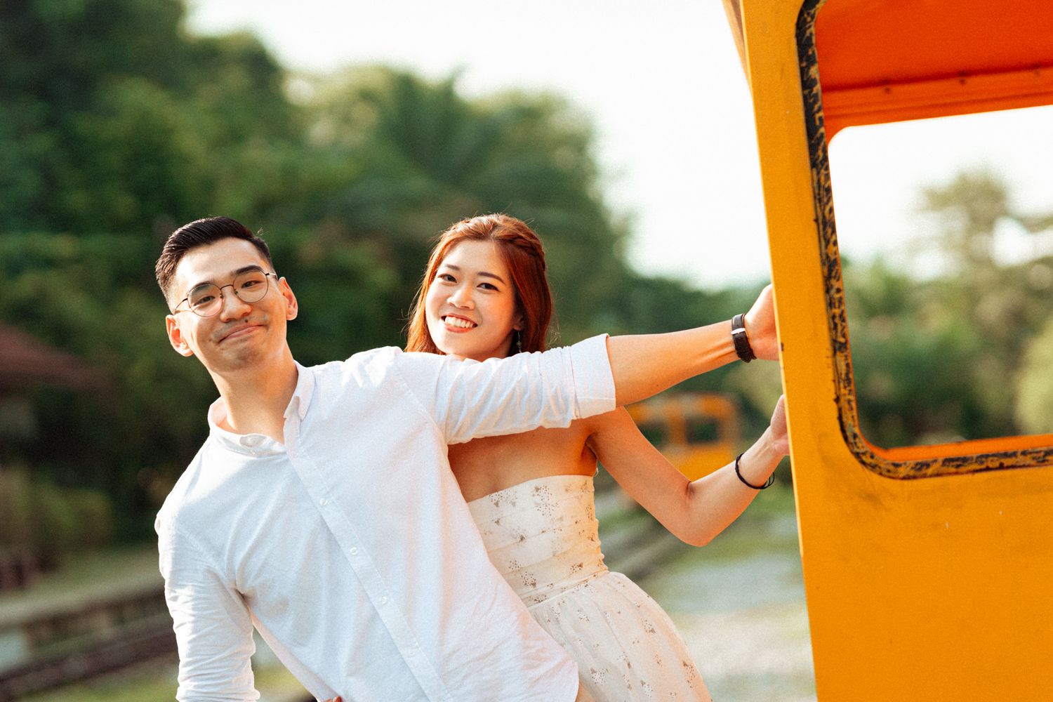 Couple in white outfits poses playfully near orange vintage train car.