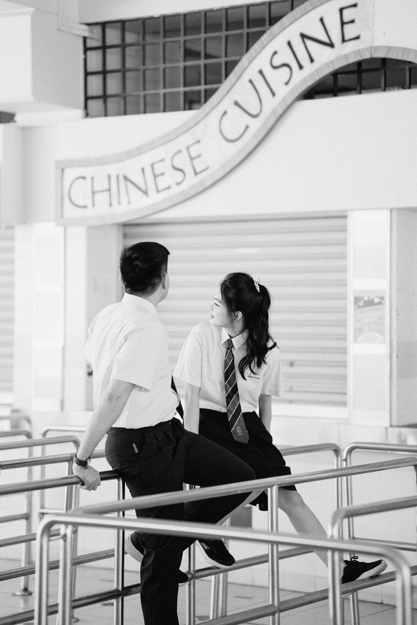 Students sitting on railings outside Chinese cuisine stalls in black and white photography.