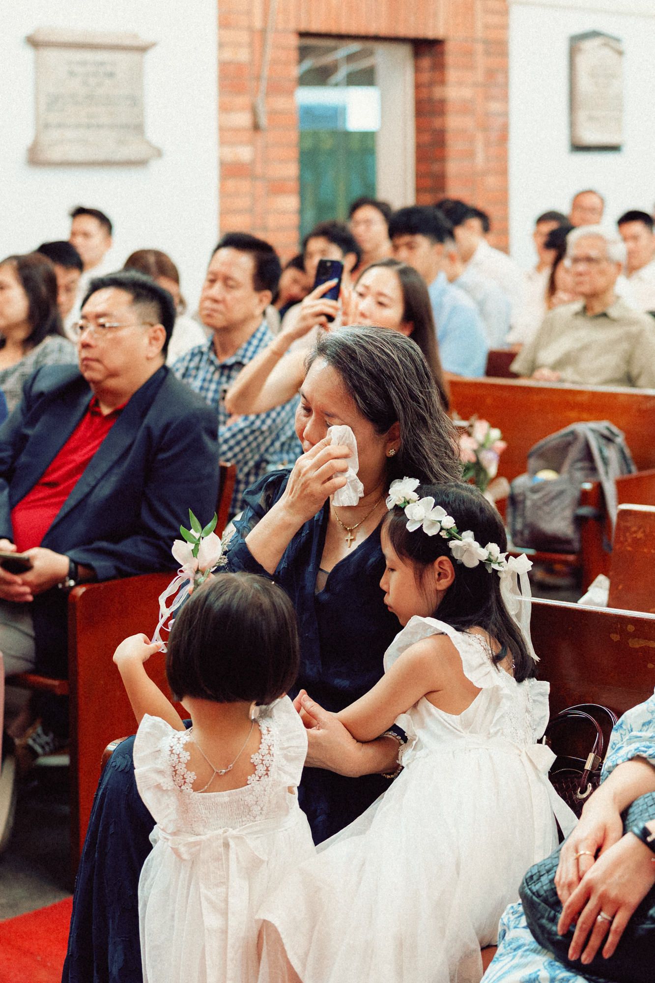 Wedding ceremony guests seated in church pews with bridesmaids wearing white dresses in the foreground.
