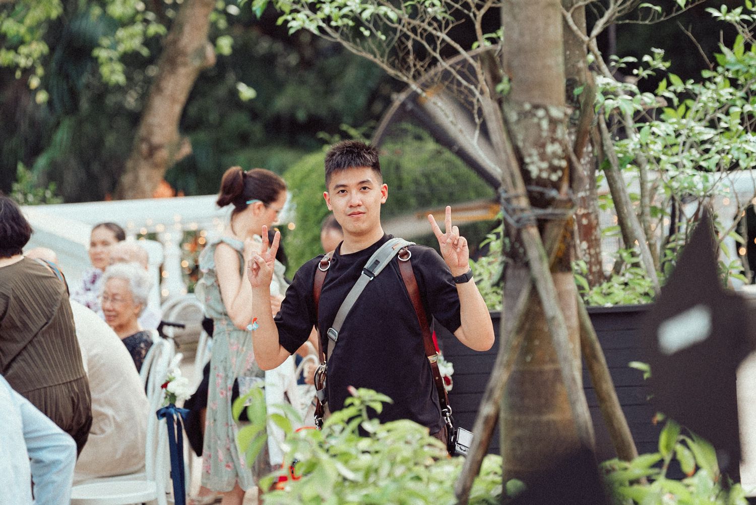 Wedding guests gather outdoors under trees during a daytime garden reception celebration.