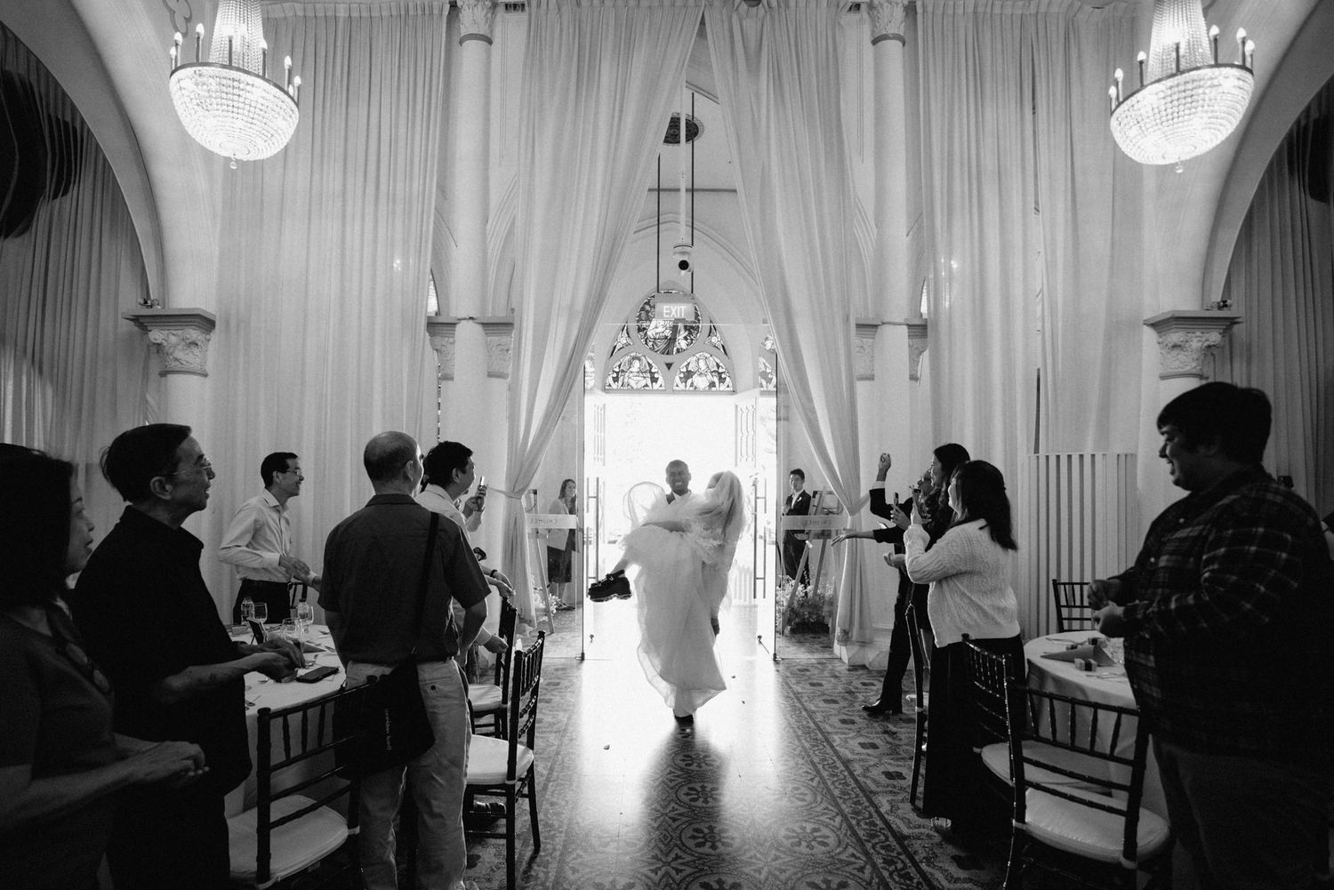 Black and white wedding ceremony photo of couple walking down aisle in elegant venue with chandeliers and draped fabric.