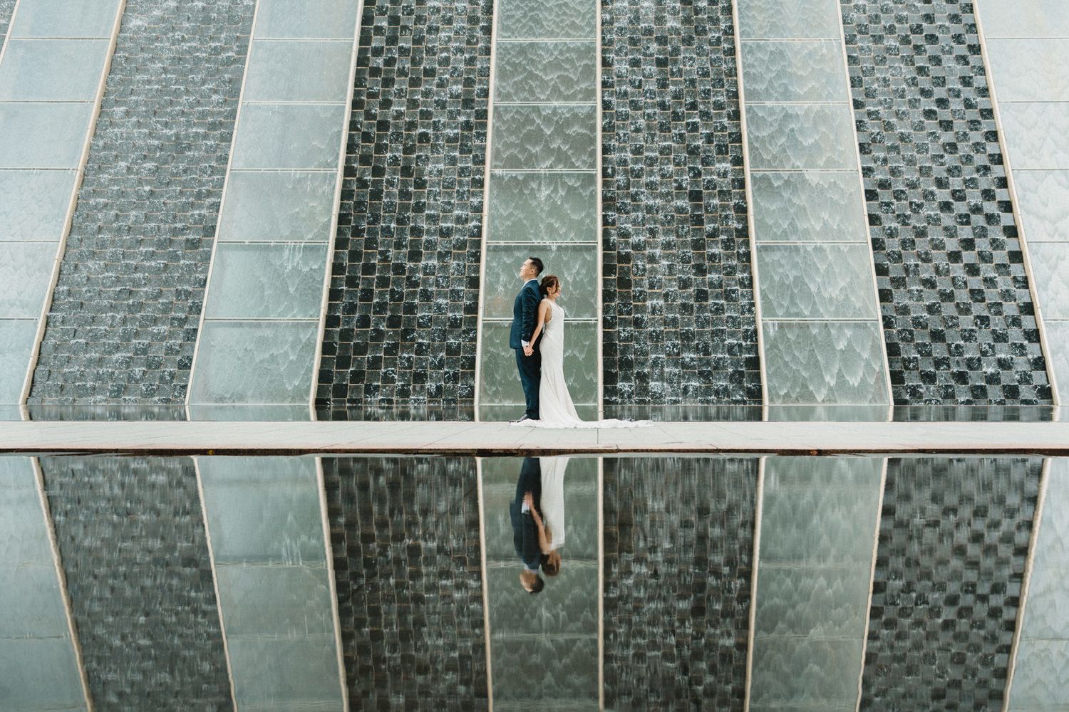 Aerial view of bride and groom standing before geometric glass wall with mirror reflection below.