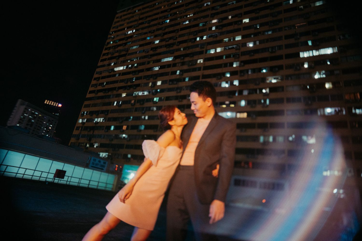 Couple posing romantically at night with illuminated city skyscraper in background.