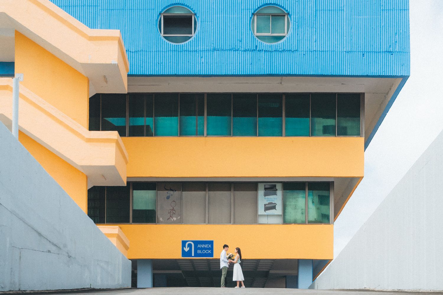 Couple embracing in front of a vibrant blue and yellow modern building with large windows.