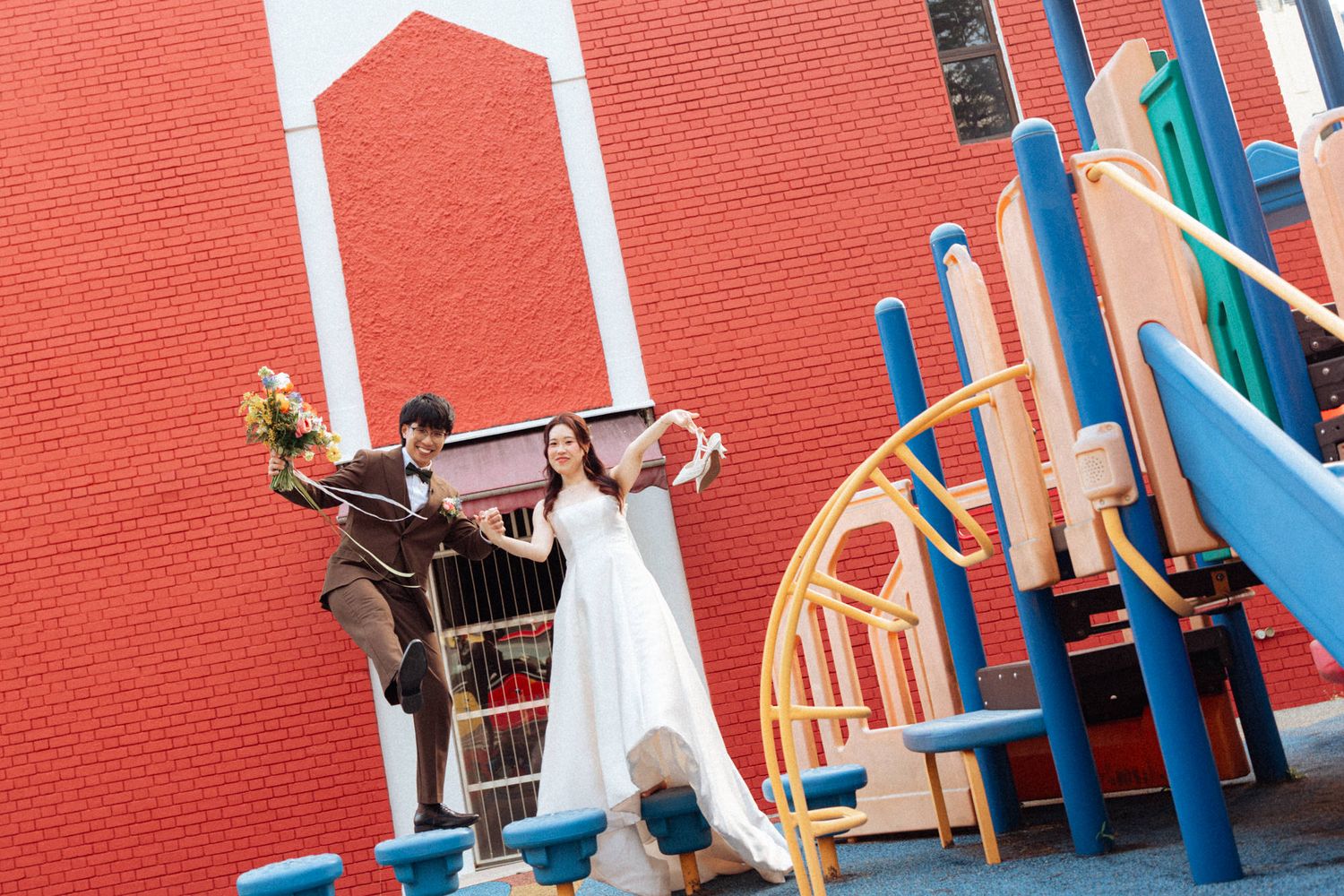 Bride and groom pose playfully at a colorful playground with a bold red brick wall backdrop.