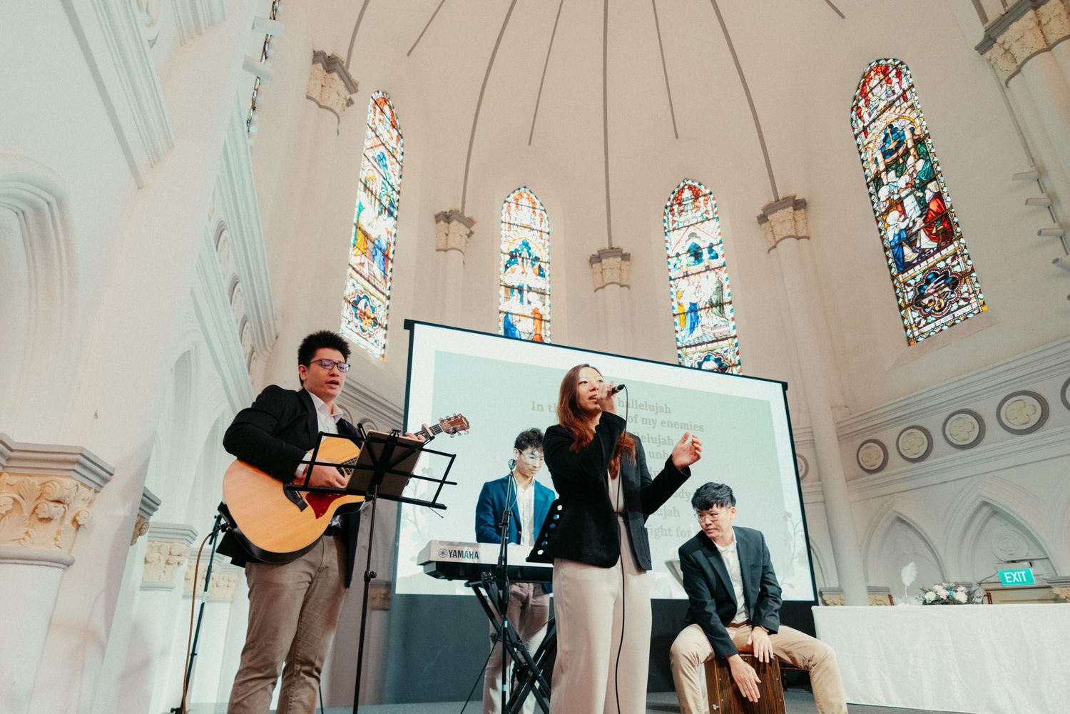 A Christian band performs in a church with stained glass windows, featuring guitar, keyboard, and vocals.