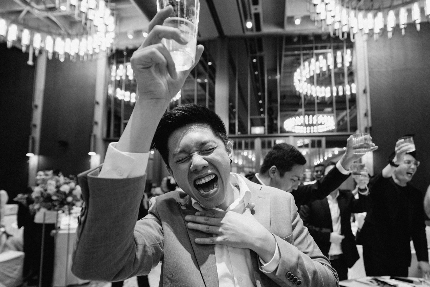 Black and white photo of a joyful man laughing and raising a glass at a formal wedding reception celebration.