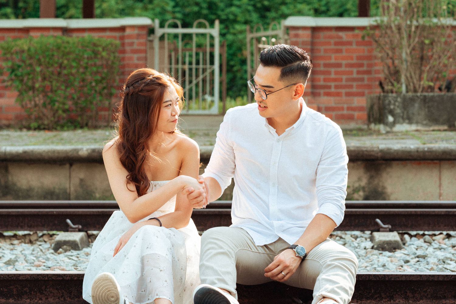 Romantic outdoor photoshoot scene with a couple in white outfits against a brick wall background.