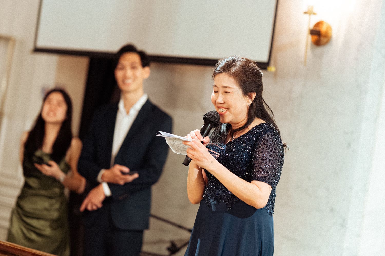 Woman in navy sequin dress speaks at microphone while couple stands behind her at formal event.