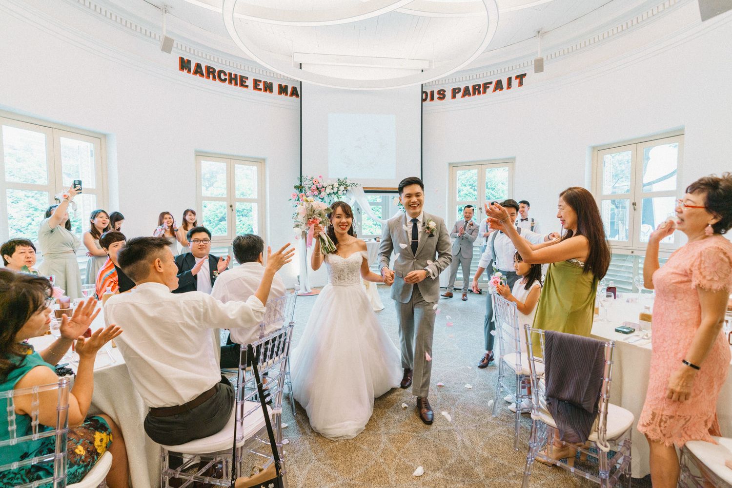 Bride and groom walk down the aisle at elegant wedding ceremony while guests applaud in a bright white venue.