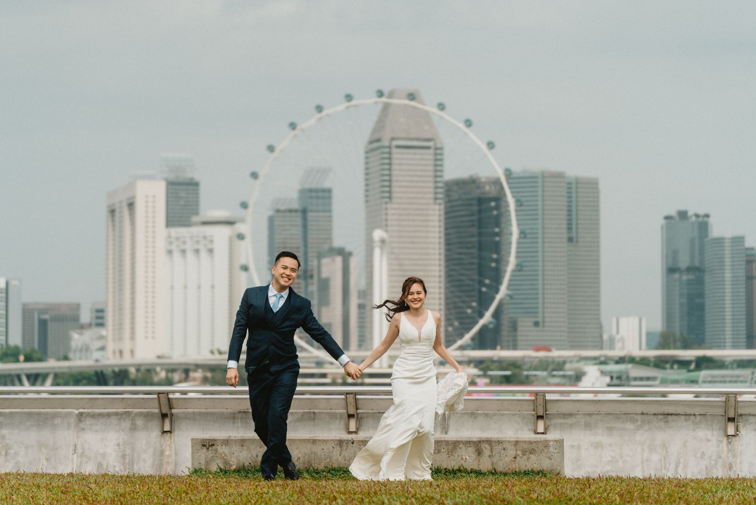 Couple in wedding attire holding hands with Singapore Flyer and city skyline in background.