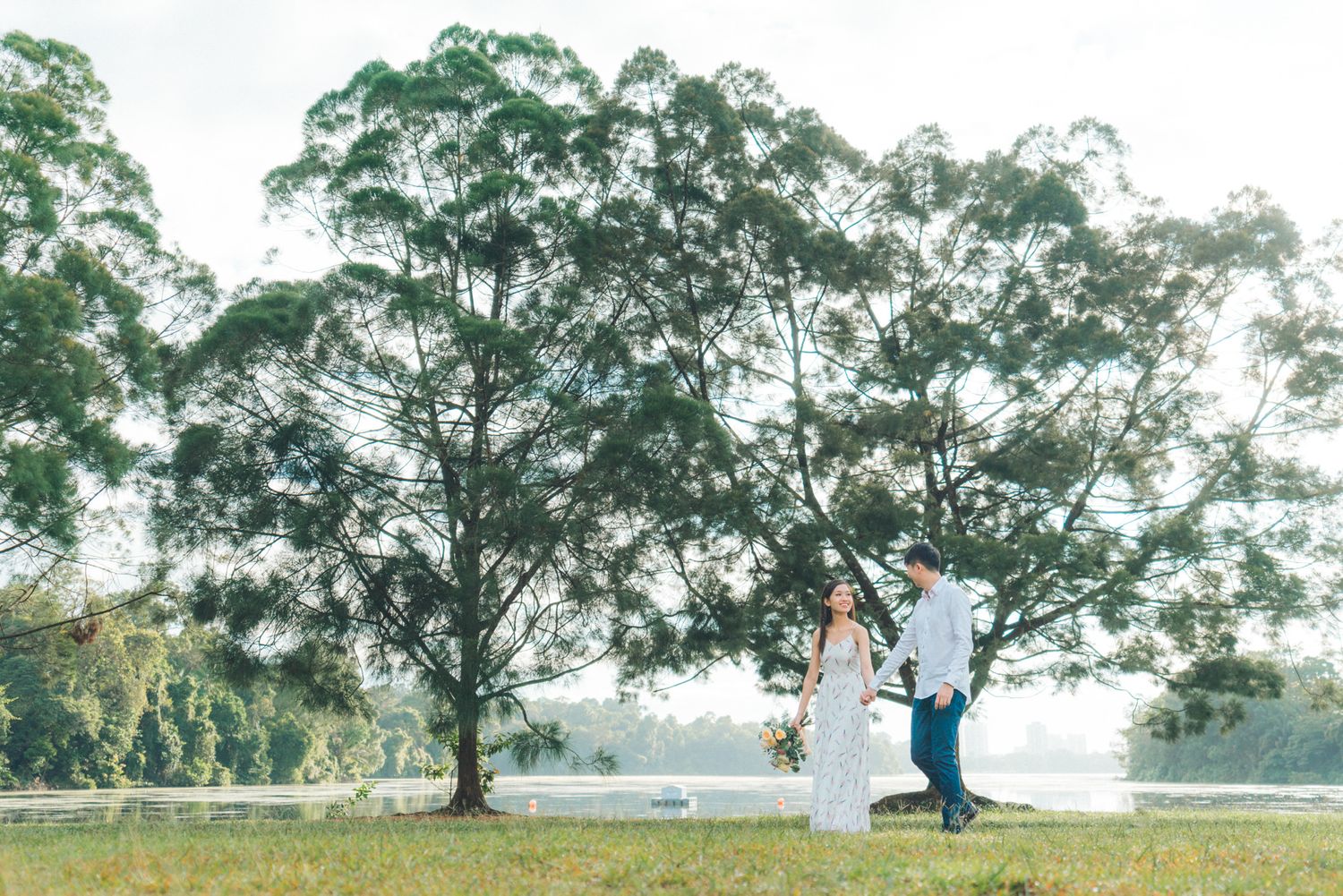 Couple standing together in a lush green park near a lake, surrounded by tall trees on a misty day.