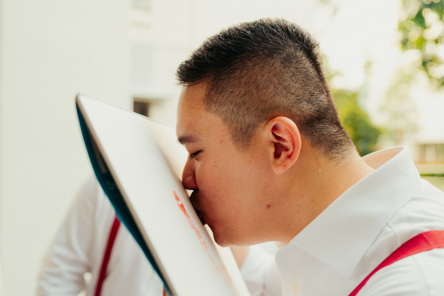 Young man in white shirt with red suspenders kissing a white book outdoors.