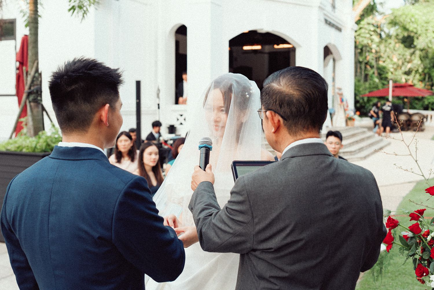 An outdoor wedding ceremony takes place in front of a white church building with guests watching from the sides.