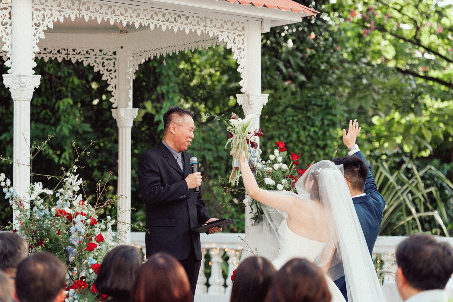 A wedding ceremony takes place under a white gazebo decorated with ornate trim and surrounded by lush gardens.
