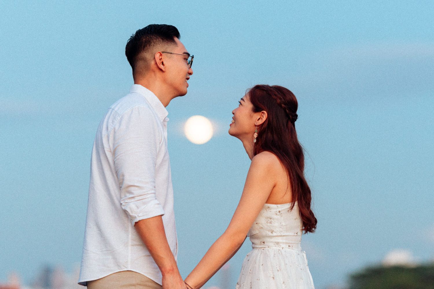 A couple in white outfits poses together against a bright blue sky during a romantic outdoor photoshoot.