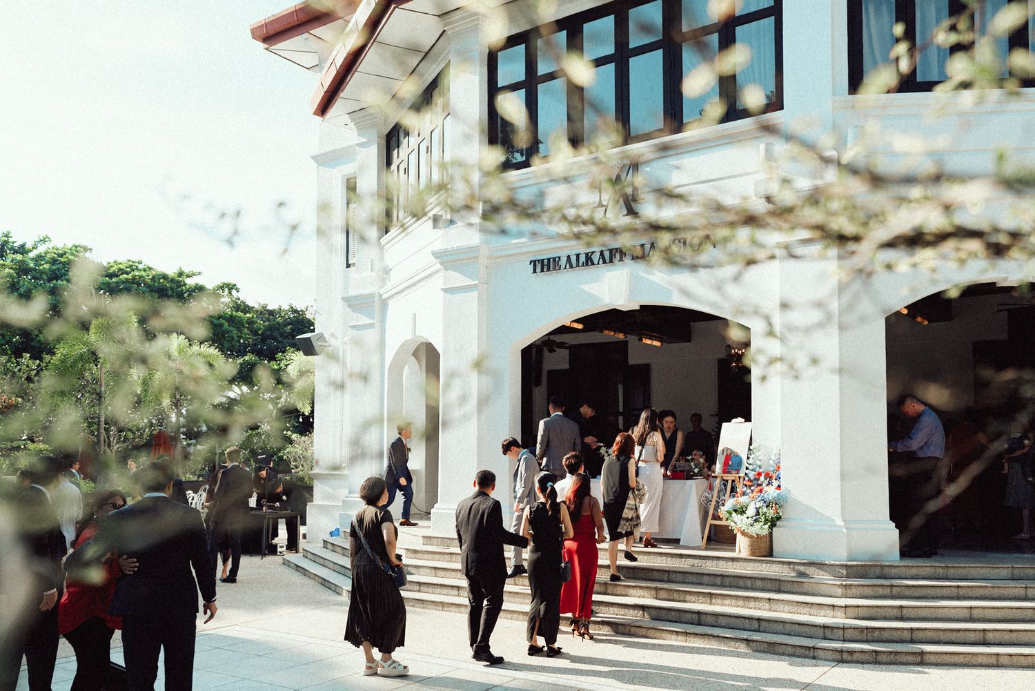 Wedding guests walking along a white Mediterranean-style building exterior with arched walkways.