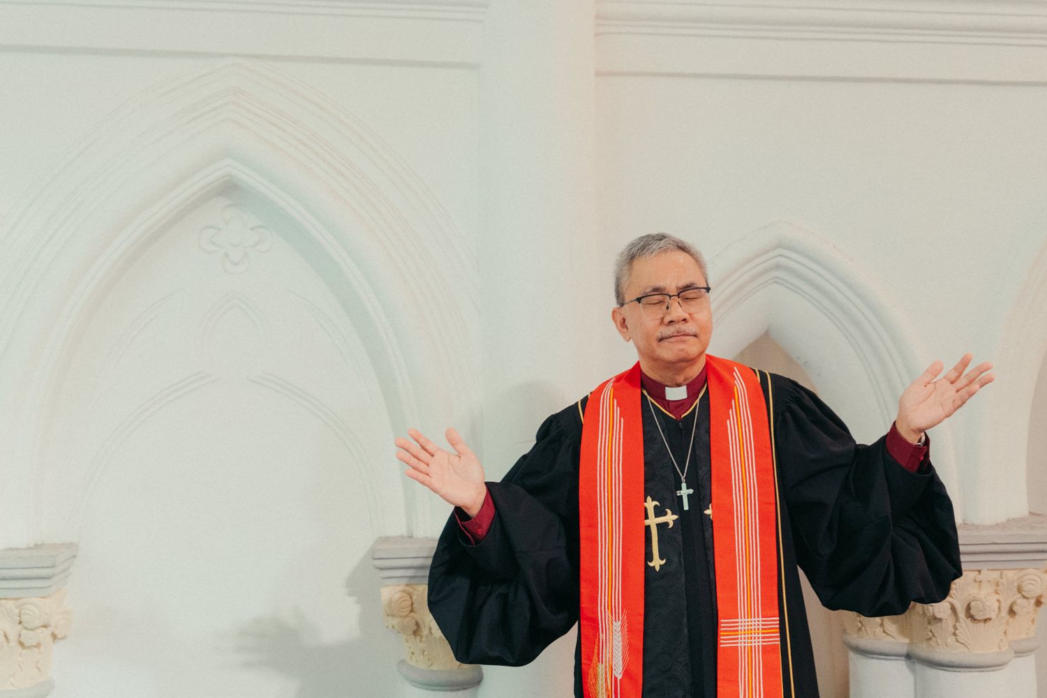 A clergy member in black robes and red stole gestures openly in a white church with Gothic arched alcoves.