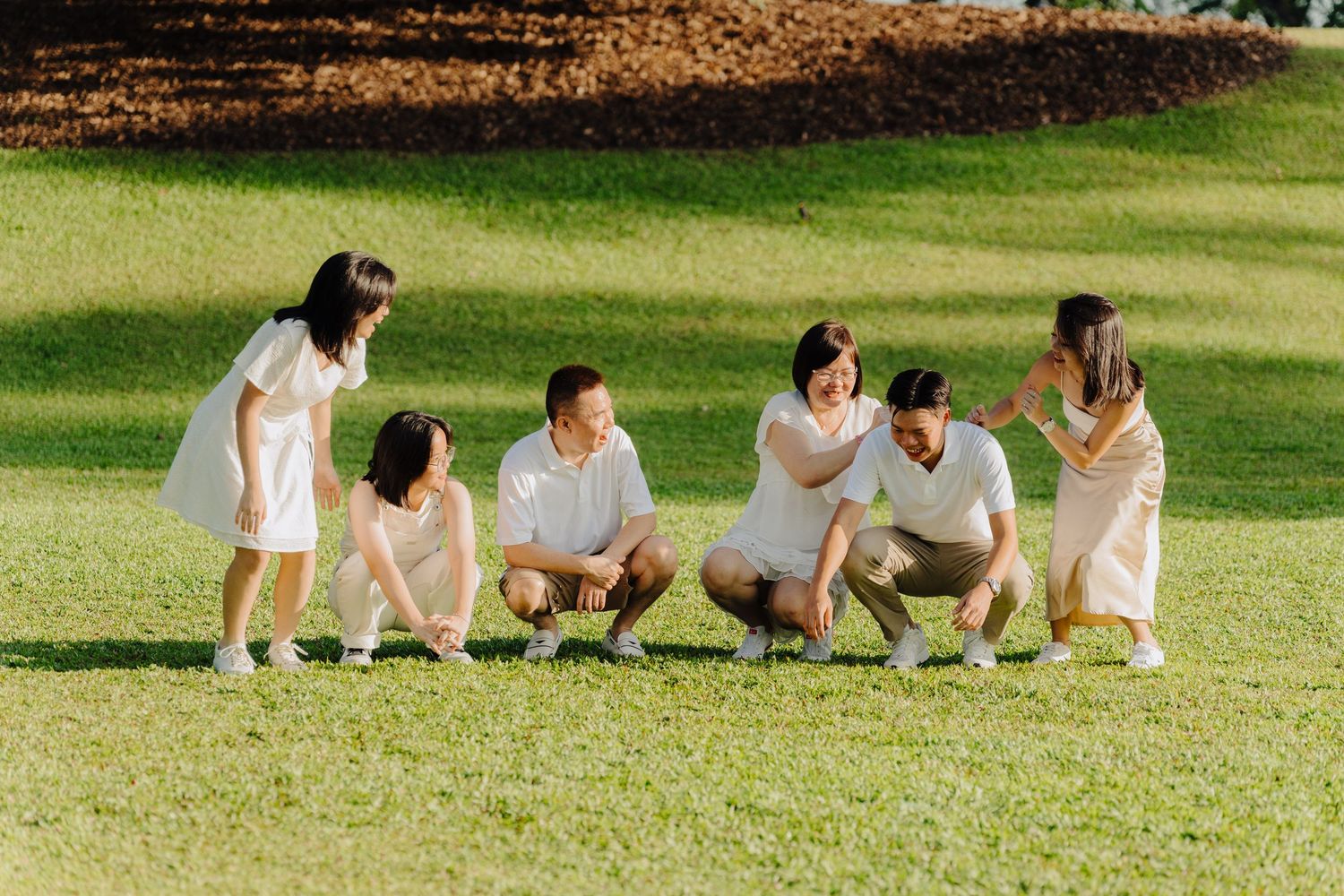 A group in white outfits sits and crouches together on a sunny grassy lawn during an outdoor photo session.