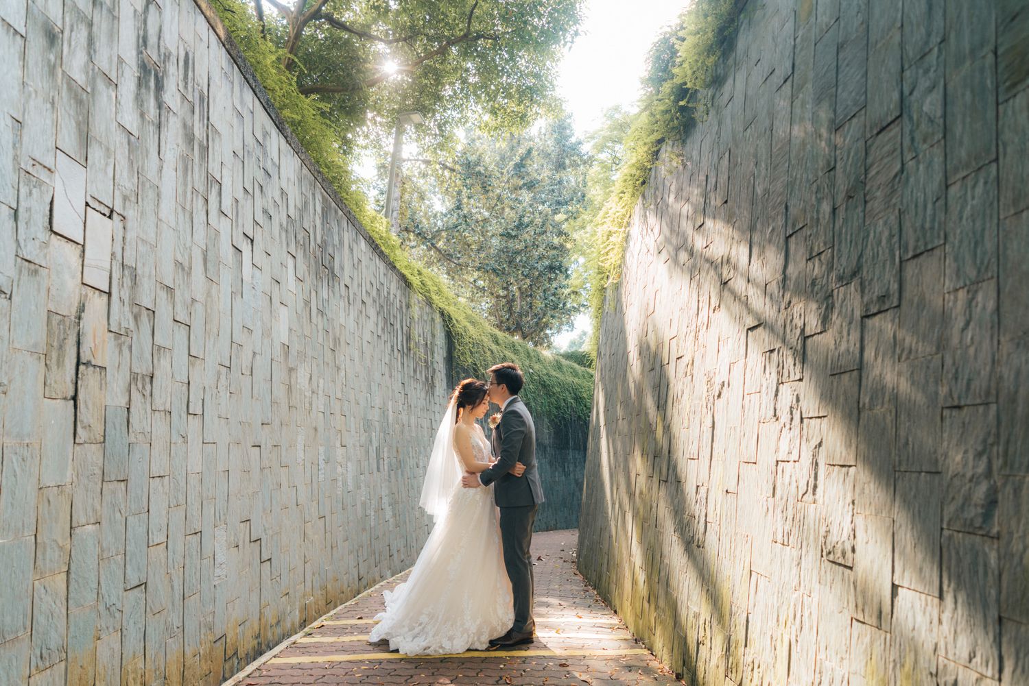 A bride and groom share a romantic moment in a sunlit stone corridor surrounded by lush greenery on their wedding day.
