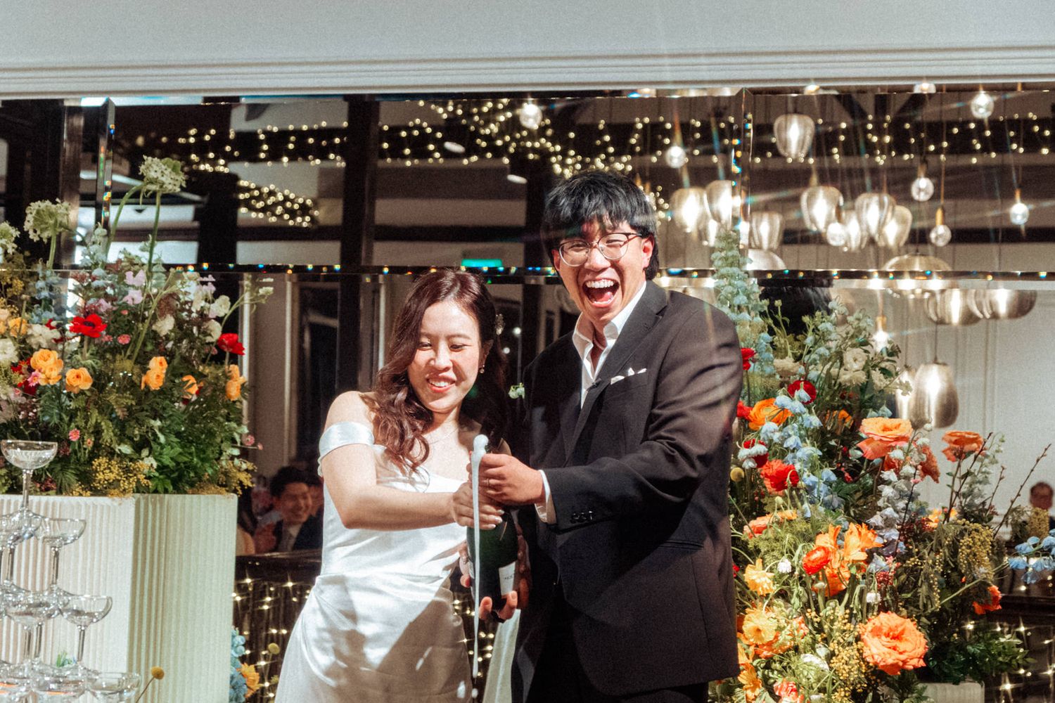 A joyful couple in wedding attire laughing together surrounded by colorful floral arrangements and warm string lights.
