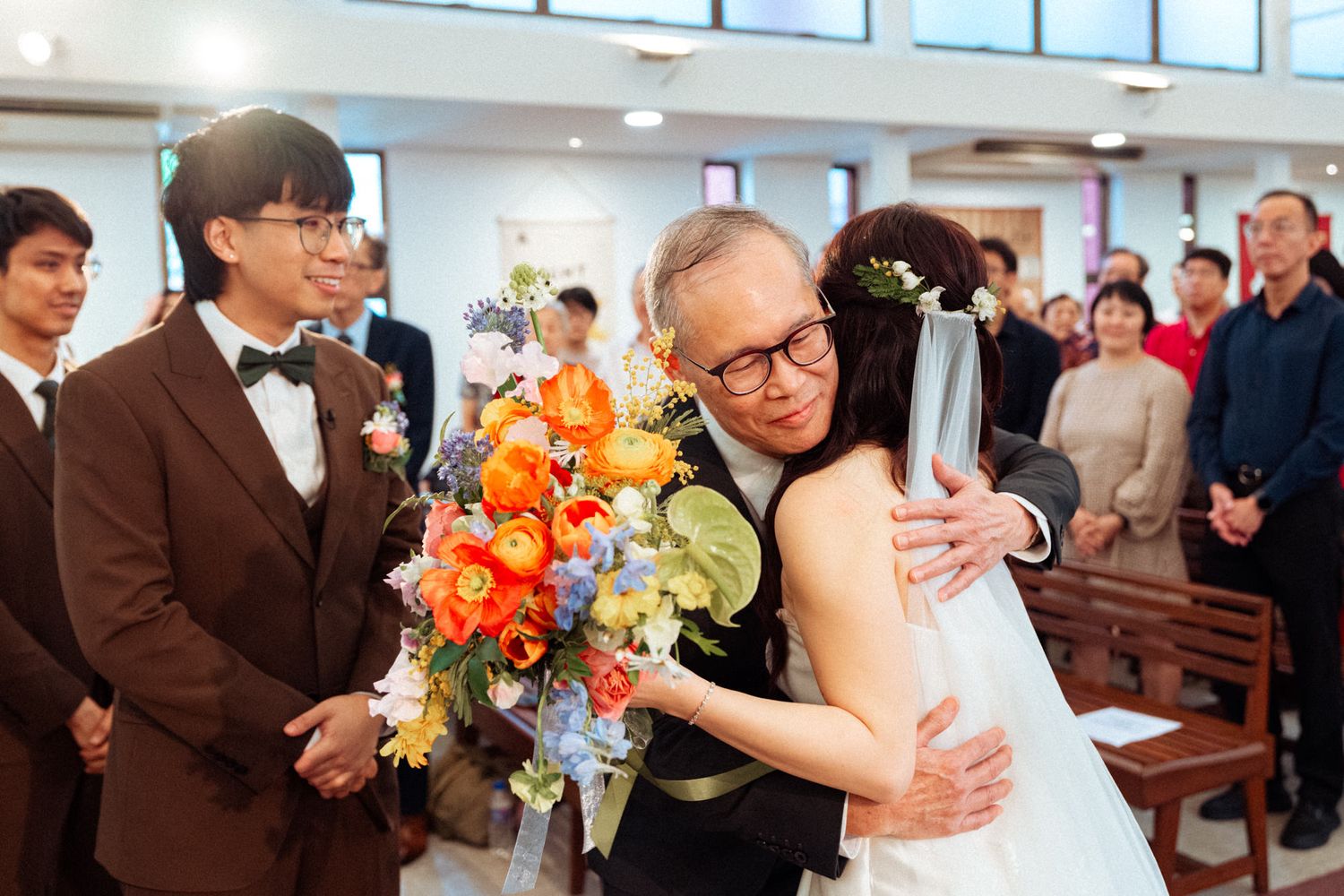 Bride in white gown hugging an older man holding colorful bouquet inside a church during a wedding ceremony.